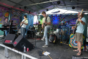 Live band performing on a stage with colorful psychedelic backdrops and a metal canopy above them, playing instruments.