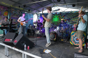 A six-member band performing on a decorated stage with psychedelic tie-dye backdrops and guitars, keyboard, and drums on display.