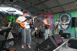 Band performing on a colorful tie-dye backdrop stage; guitarist in a white shirt in the foreground, two other guitarists and a drummer in the back.