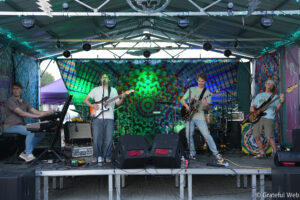Band performs on a colorful tie-dye stage backdrop with guitars, keyboards, and a drummer visible in the background.
