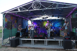Live band performing on an outdoor stage with psychedelic tie-dye backdrops and purple lighting.