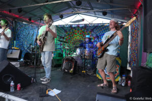 Band performing on a colorful tie-dye stage backdrop; lead singer with guitar at center, bassist to the right, drummer in the back left/right.