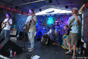 Live band performing on a colorful tie-dye stage backdrop, with guitarist left, singer center, bassist right, and drummer in the back.