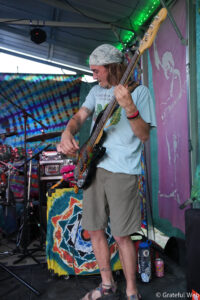 Bass guitarist wearing a bandana and light blue shirt plays a bass on a colorful tie-dye stage backdrop during a live performance.
