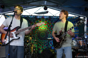 Two guitarists perform on a colorful tie-dye stage backdrop, one singing into a microphone while playing a sunburst Stratocaster; the other plays a dark guitar.
