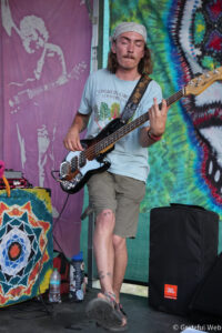 Bass guitarist performing on a colorful stage with tie-dye backdrop, wearing a light blue shirt and bandana.