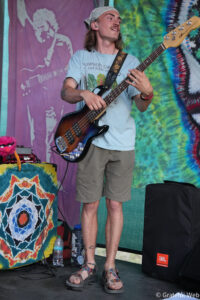 Male bassist on a colorful tie-dye stage backdrop playing a sunburst bass guitar with a decorated strap.
