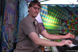 Man with glasses playing a keyboard, performing in a colorful festival booth with tie-dye banners behind him.