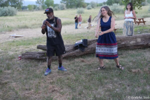 Man in a sleeveless black shirt and cap dances near a fallen log in an open field while a woman in a blue patterned dress watches, with others in the background.