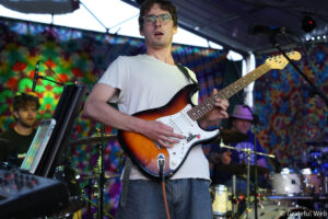 Male guitarist with glasses in a white shirt playing a sunburst Fender Stratocaster on a colorful tie-dye stage backdrop.