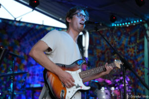 Male guitarist singing into a microphone on a colorful, lit stage backdrop.
