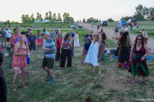 Group of people dancing and socializing at an outdoor festival on a grassy field, wearing colorful bohemian clothing at dusk.