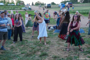 Group of adults dancing and socializing on a grassy field at an outdoor festival, wearing colorful outfits and dresses.