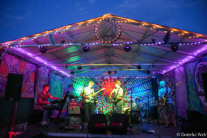 Band performs on a brightly lit stage with tie-dye backdrops and string lights at a festival venue.