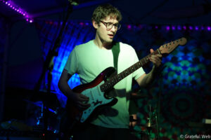 Male guitarist wearing glasses and a light-colored T-shirt plays a red Fender Stratocaster on a dimly lit stage with purple/blue lighting and a blurred crowd in the background.