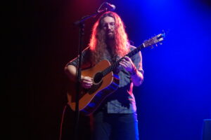Musician with long hair and glasses playing an acoustic guitar on stage under red and blue lights.