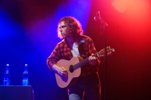 Male guitarist in a red plaid shirt playing an acoustic guitar on stage with blue and red lighting.