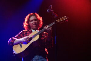 Guitarist on stage wearing a red plaid shirt, playing an acoustic guitar under blue and red stage lighting.