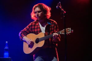 Male musician in a red plaid shirt playing an acoustic guitar on a dark stage with blue and red lighting