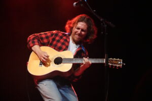 Male guitarist playing an acoustic guitar on stage, wearing a red plaid shirt and jeans, intense expression, microphone in front.