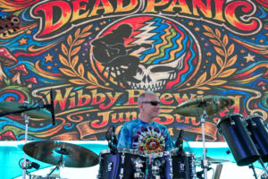 Drummer wearing sunglasses plays a drum kit on a stage with a bright psychedelic backdrop featuring a skull design and swirls of colored art.