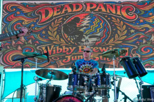 Drummer on stage with a colorful psychedelic backdrop and 'Deadpan' banner, guitarist visible on the left.