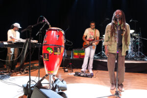 Band on stage performing live: vocalist with dreadlocks at the mic, guitarist, and keyboardist with a red conga drum in the foreground; drums and cables visible in back.