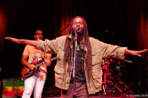 Lead singer with long dreadlocks and a khaki jacket, arms outstretched, singing into a microphone on stage. Background guitarist and drummer under red stage lights.