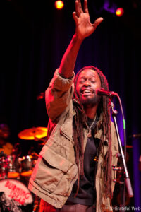 Male singer with long dreadlocks raises his hand on stage, microphone in front, wearing a tan jacket under red stage lights.