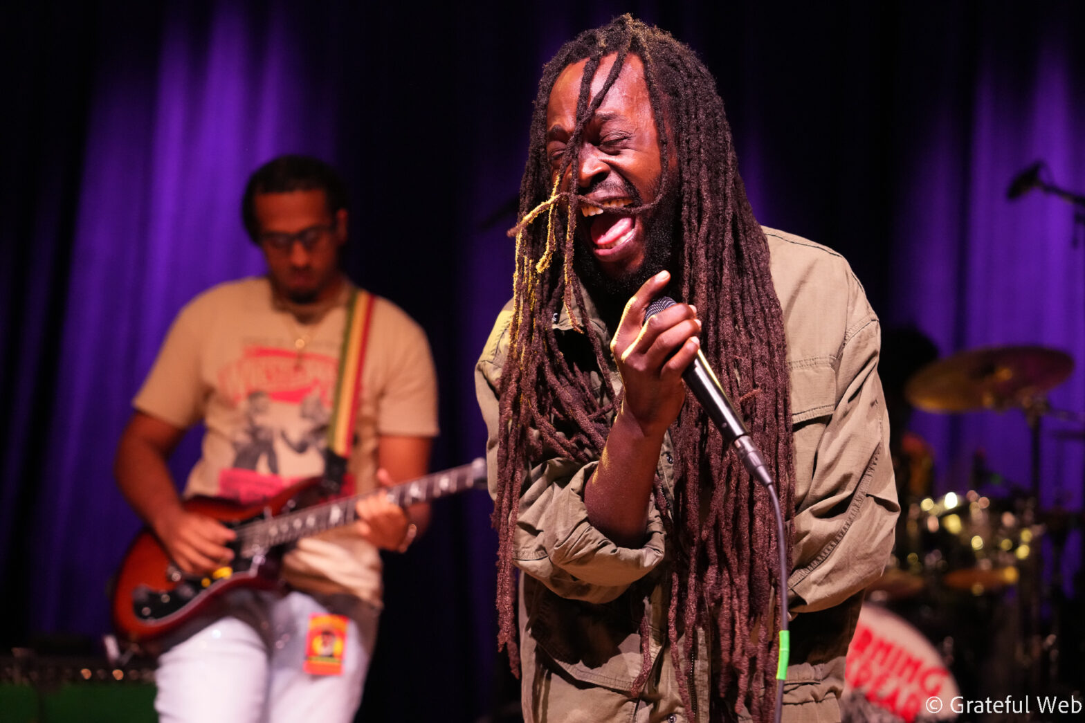 Singer with long dreadlocks passionately singing into a microphone on a purple-lit stage, guitarist in the background.