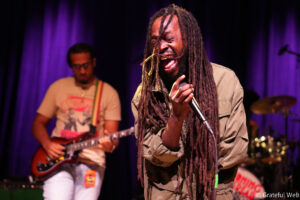 Singer with long dreadlocks passionately singing into a microphone on a purple-lit stage, guitarist in the background.