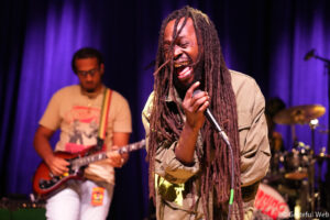 Male vocalist with long dreadlocks singing into a microphone on a dimly lit stage; guitarist in the background under purple lights.