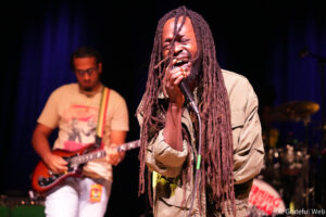 Male singer with long dreadlocks passionately singing into a microphone on stage, wearing a beige shirt; guitarist blurred in the background.