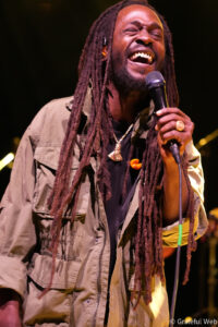 Male singer with long dreadlocks performing on stage, gripping a microphone and laughing into it, wearing a beige jacket and jewelry.