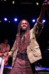 Male performer with long dreadlocks on stage, raising a mic/hand toward the crowd as blue stage lights shine behind him.