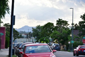 Busy urban street lined with parked cars, trees, and a distant hill under a cloudy sky; signs and a stop sign visible on the right side.