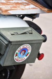 Green metal storage box on the rear of a motorcycle, featuring a Grateful Dead sticker with a skull and red roses.