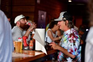 Two men at a wooden bar: one in a white cap drinks from a red can, the other in a tie-dye shirt and gray cap smiles while looking at a phone/device on the counter.