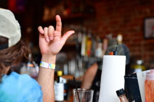 Bar patron at a dimly lit bar raises a hand in a shaka (hang loose) gesture, thumb and pinky extended.