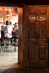 Open wooden door reveals a busy bar interior with patrons seated on stools and shelves of bottles in the background.