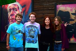 Four people posing and smiling in front of a dark wooden wall with colorful murals behind them.