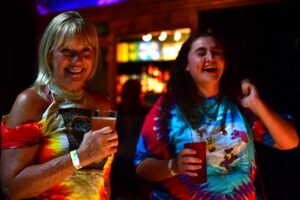 Two women laughing with drinks at a dimly lit bar or party venue in the background.