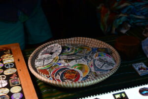 Basket of colorful round stickers and pins on a market table, with 'Please be kind' visible among the designs.