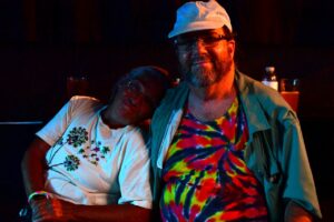 Two friends posing together at a dimly lit event; woman in a white shirt with floral embroidery leans on a man in a tie‑dye shirt and cap.