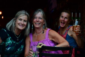 Three smiling women at a bar posing for the camera, each with a drink and leaning together.