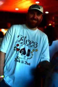 Smiling man wearing a cap and a white T-shirt with a playing-card bouquet graphic reading 'Ace Back to Back' in a warm, dim venue.