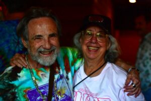 Two smiling seniors with arms around each other at a colorful party.