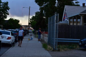 Sidewalk scene at dusk with a man walking, a child playing with a toy, and parked cars along the street (fence and house on the right).
