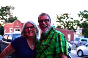 Smiling couple wearing glasses posing outdoors on a street with cars and brick buildings in the background.