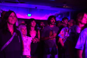 Group of people standing in a dim venue under purple stage lights, watching a performance attentively.